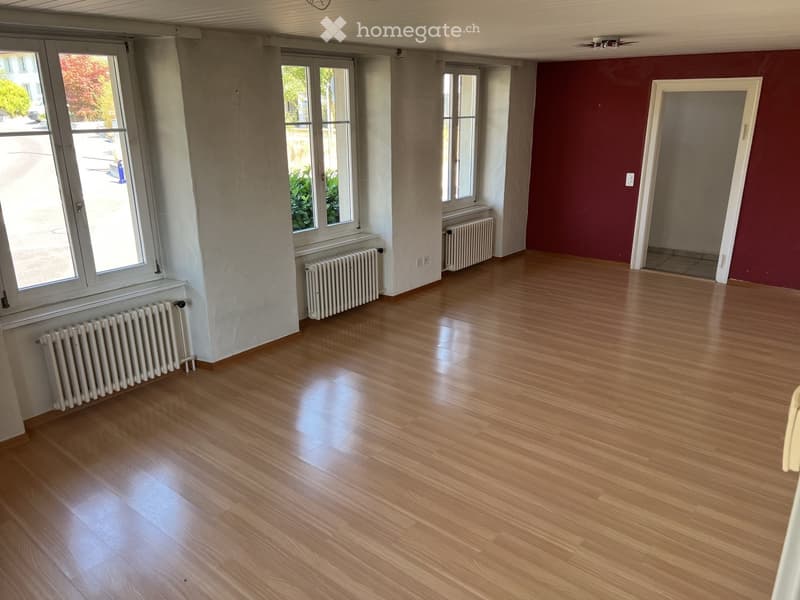 Empty room with wooden flooring, white radiators beneath windows, and a red accent wall. No furniture or equipment present.