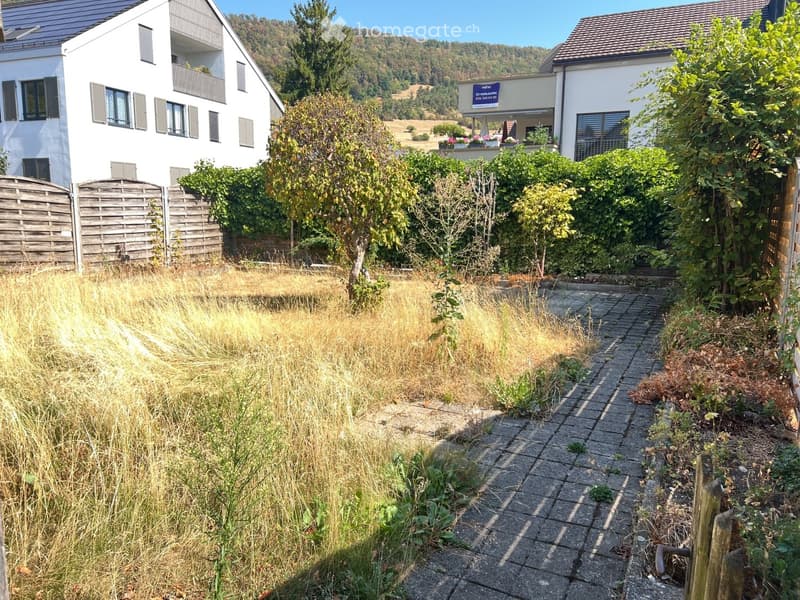 Garden area with a paved pathway, overgrown grass, and surrounding greenery, with two modern houses in the background.