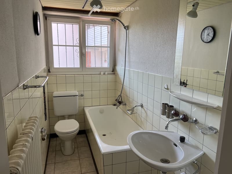 Bathroom with a toilet, bathtub, sink, radiator, mirror, and clock. The walls are tiled, and there is a window with bars.