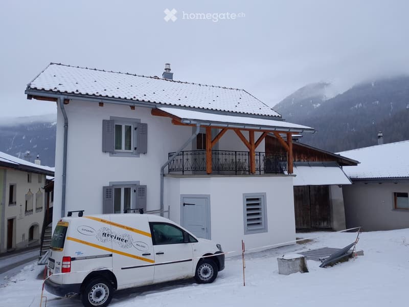 Two-story white building with grey shutters, snow-covered roof, and balcony with metal railing. A white van is parked in front.