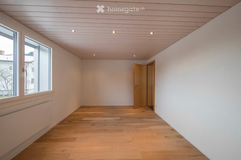 Empty room with light wooden flooring, white walls, and a wooden door leading to another space. Large windows allow natural light to enter.