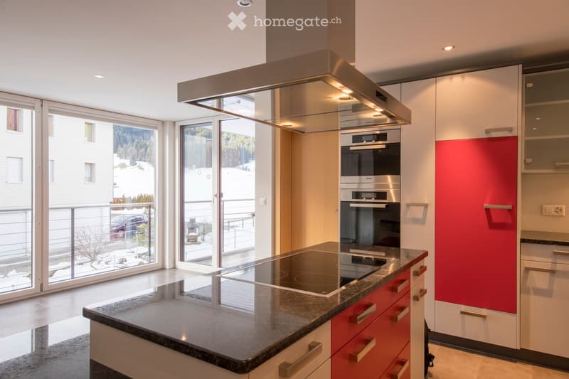 Modern kitchen with white and red cabinets, granite countertop, induction stove, built-in oven, and range hood. Large windows offer a view of the outdoor area.