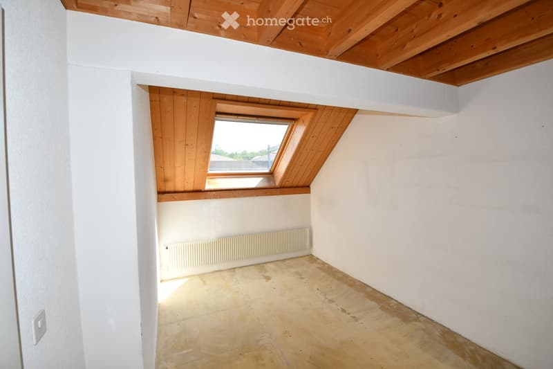 Empty room with white walls, wooden ceiling beams, and a skylight window. The floor is unfinished, and there is a radiator on one wall.