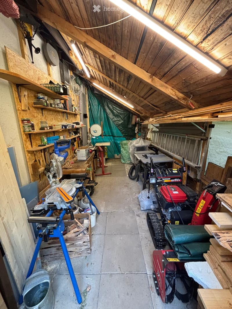 Workshop with exposed wooden beams, concrete floor, and various tools and equipment including a saw, table, shelves, and a lawn mower.