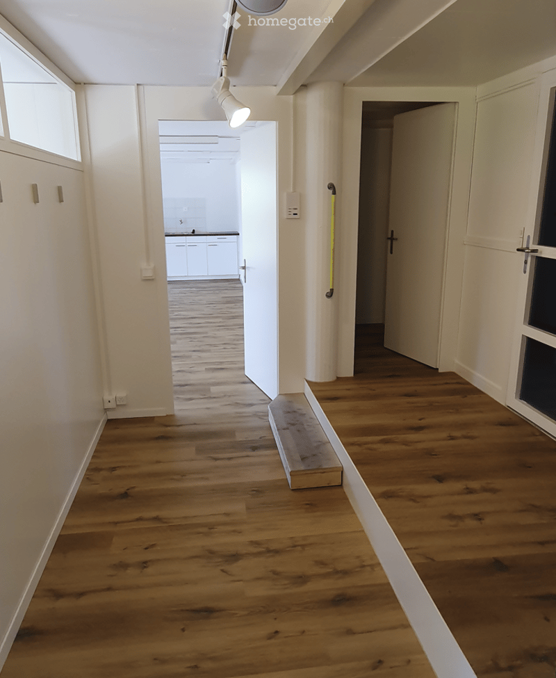 Empty hallway with wooden flooring, white walls, and a small bench placed near the wall. The hallway leads to an open doorway revealing a kitchen area with white cabinets and a countertop.