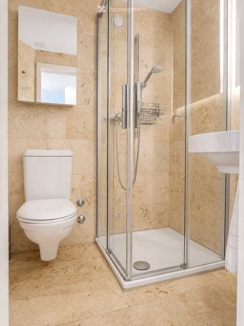 Modern bathroom with beige tiled walls and floor, featuring a white toilet, a glass-enclosed shower with a handheld showerhead, a white sink, and a mirrored cabinet above the sink.