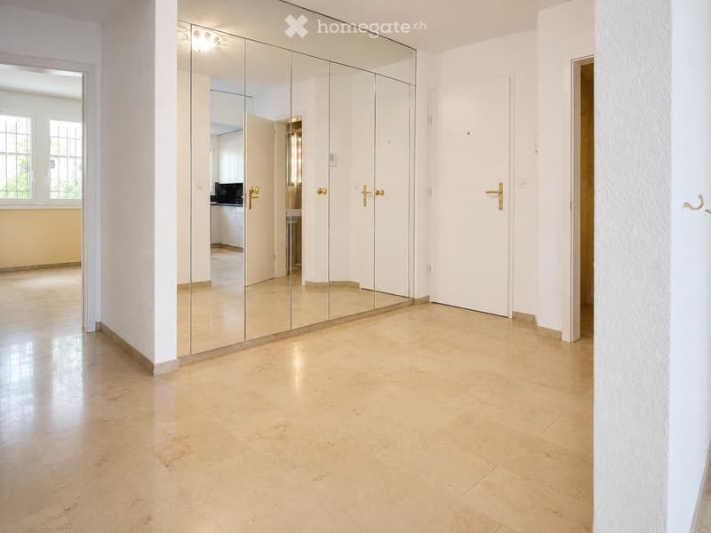 Bright hallway with beige tiled flooring, white walls, and large mirrored cabinet. Features multiple doors and a window on the left side.