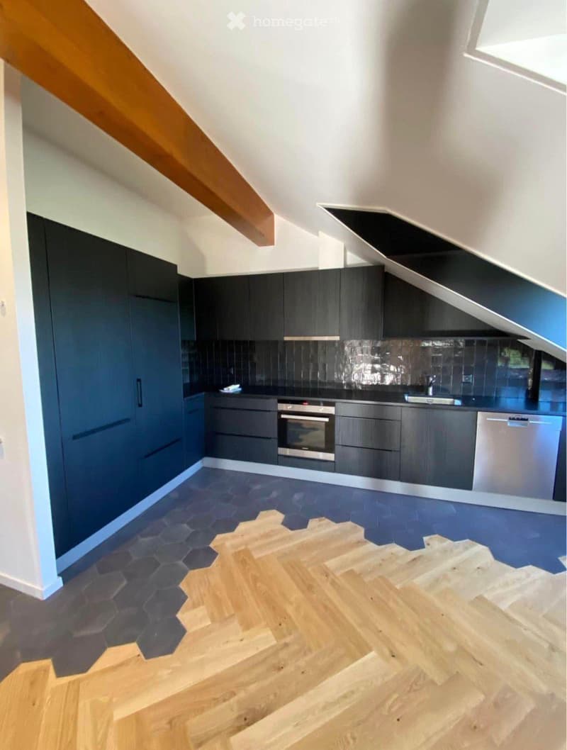 Modern kitchen with dark grey cabinets, stainless steel appliances, and a sleek countertop. The floor features a mix of hexagonal tiles and wooden planks, with a wooden staircase leading upwards.