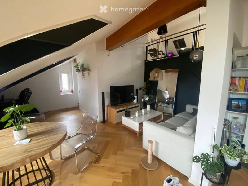 Modern living room with wooden flooring, white walls, and a slanted ceiling. Features include a white sofa, a coffee table, a dining table with chairs, a cat tree, and several plants. A TV is mounted on a wooden stand, and a large mirror is positioned on