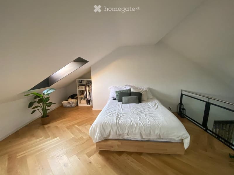 Cozy attic bedroom with wooden flooring, a large bed with white bedding and multiple pillows, a wardrobe, a potted plant, and a lantern on the floor near a glass railing.