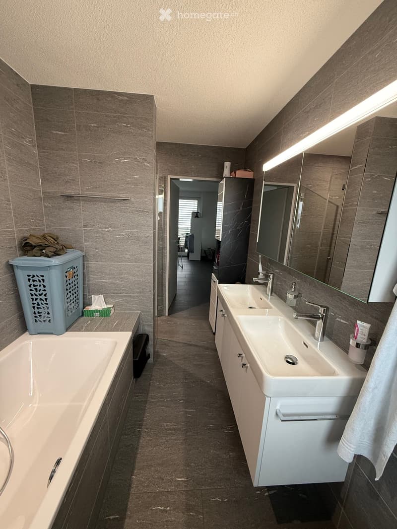 Modern bathroom with a double sink vanity, bathtub, large mirror, and grey tiled walls and floor. Laundry basket and trash bin are visible.