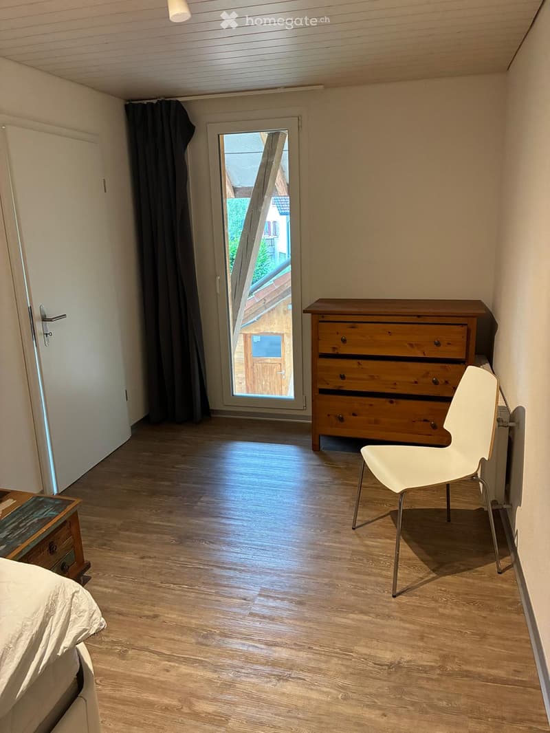 Bright bedroom with wooden flooring, a bed with white bedding, a white chair, and a wooden chest of drawers. A large window with a view of the outside is partially covered by dark curtains.