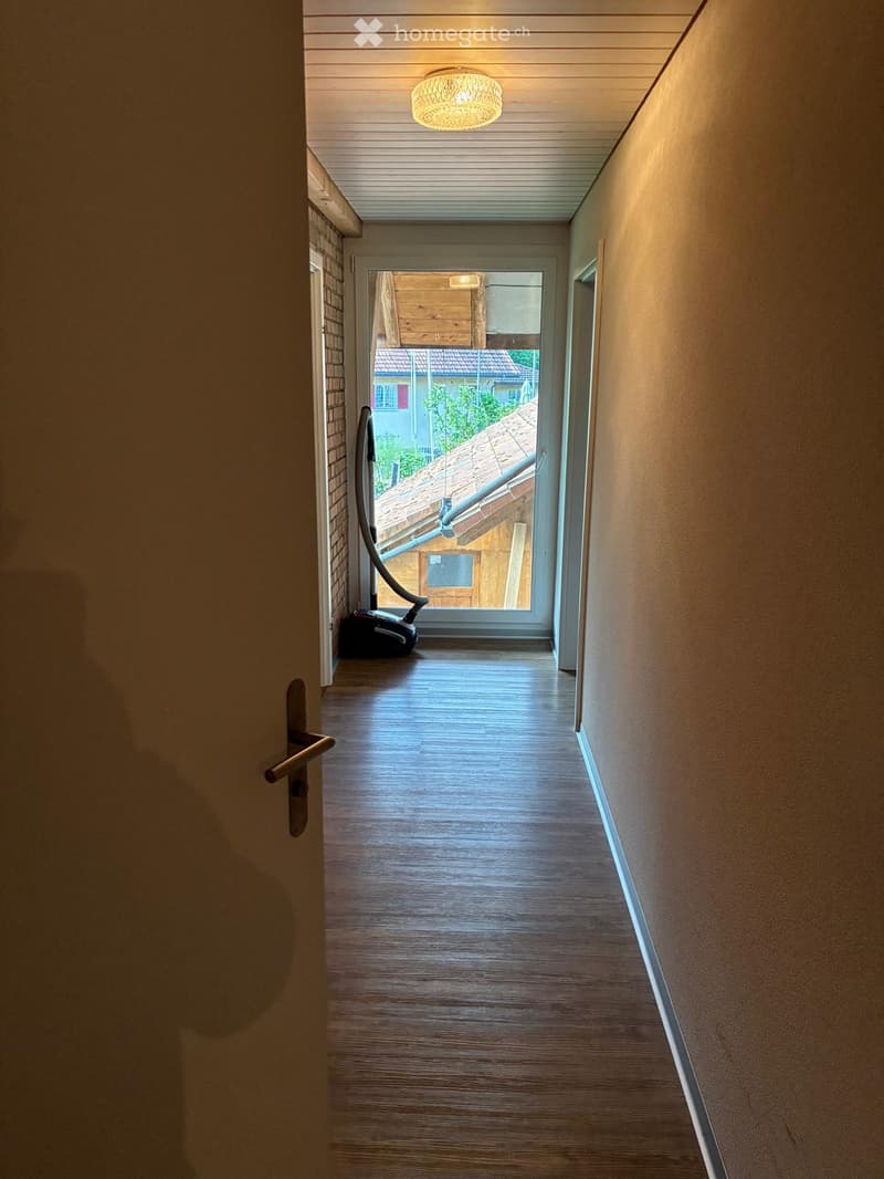 A narrow hallway with wooden flooring, beige walls, and a ceiling light fixture. A vacuum cleaner is placed against the left wall, and a door is visible on the left side.