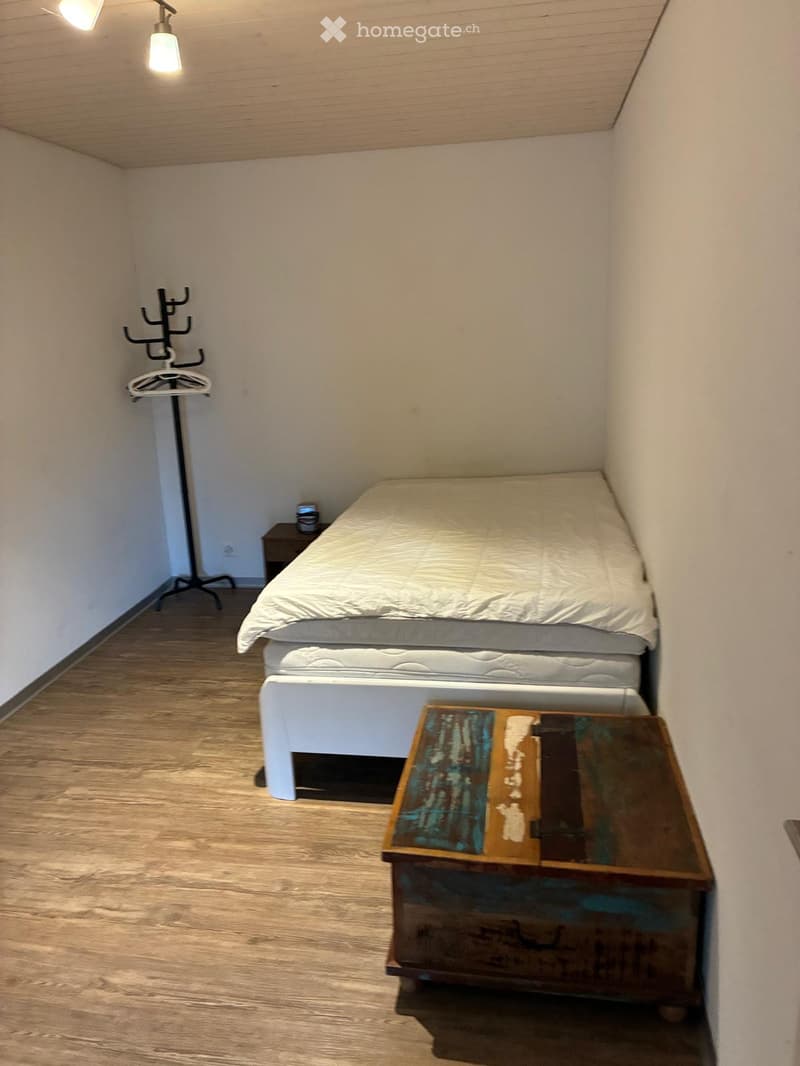Small bedroom with a single bed, nightstand, and coat rack. Light wooden flooring and white walls.