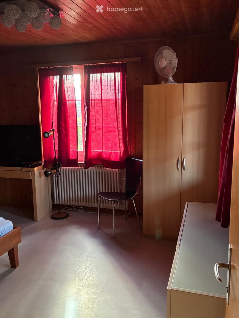 Cozy bedroom with wooden walls, a bed, a chair, a wardrobe, a fan on top, a table with a lamp, and red curtains on the window.