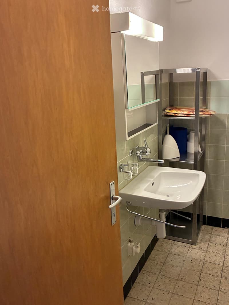 Modern bathroom with a white sink, chrome faucet, mirror cabinet, and a toilet in the corner. The walls are tiled in a light green color, and the floor has beige tiles.