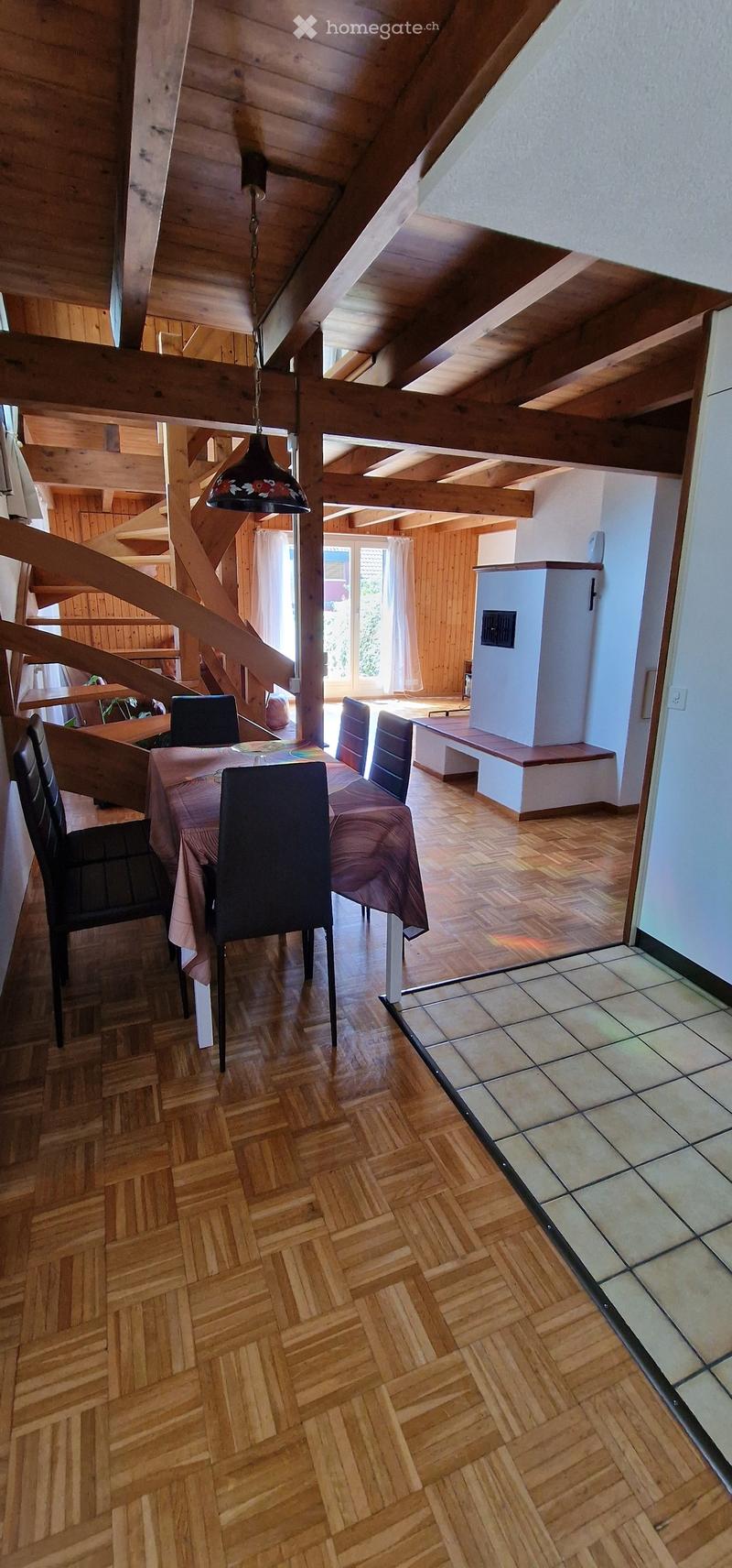 Dining area with wooden herringbone flooring, a dining table with chairs, a bench, and a refrigerator in the background. The ceiling features exposed wooden beams and a hanging light fixture.