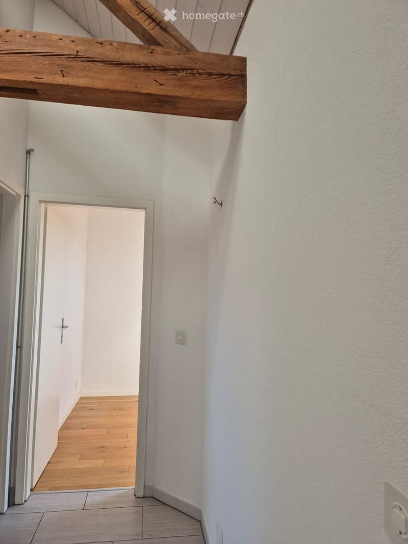 Empty hallway with white walls, wooden floor, and a wooden beam on the ceiling. A doorway leads to another room with wooden flooring.