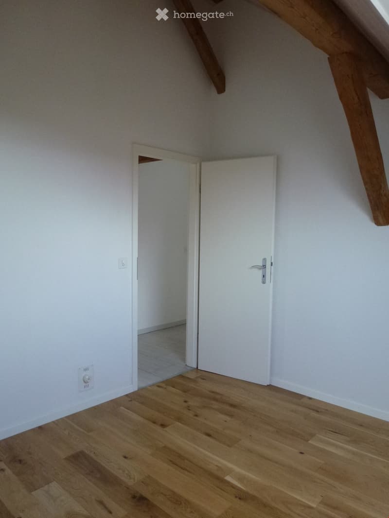 Empty attic room with wooden flooring, white walls, and exposed wooden beams. Features a closed white door and an open doorway leading to another space.