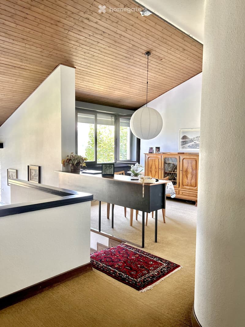 Modern living room with a slanted wooden ceiling, large windows, a round ball lamp, a wooden cabinet, a table with chairs, and a patterned rug.