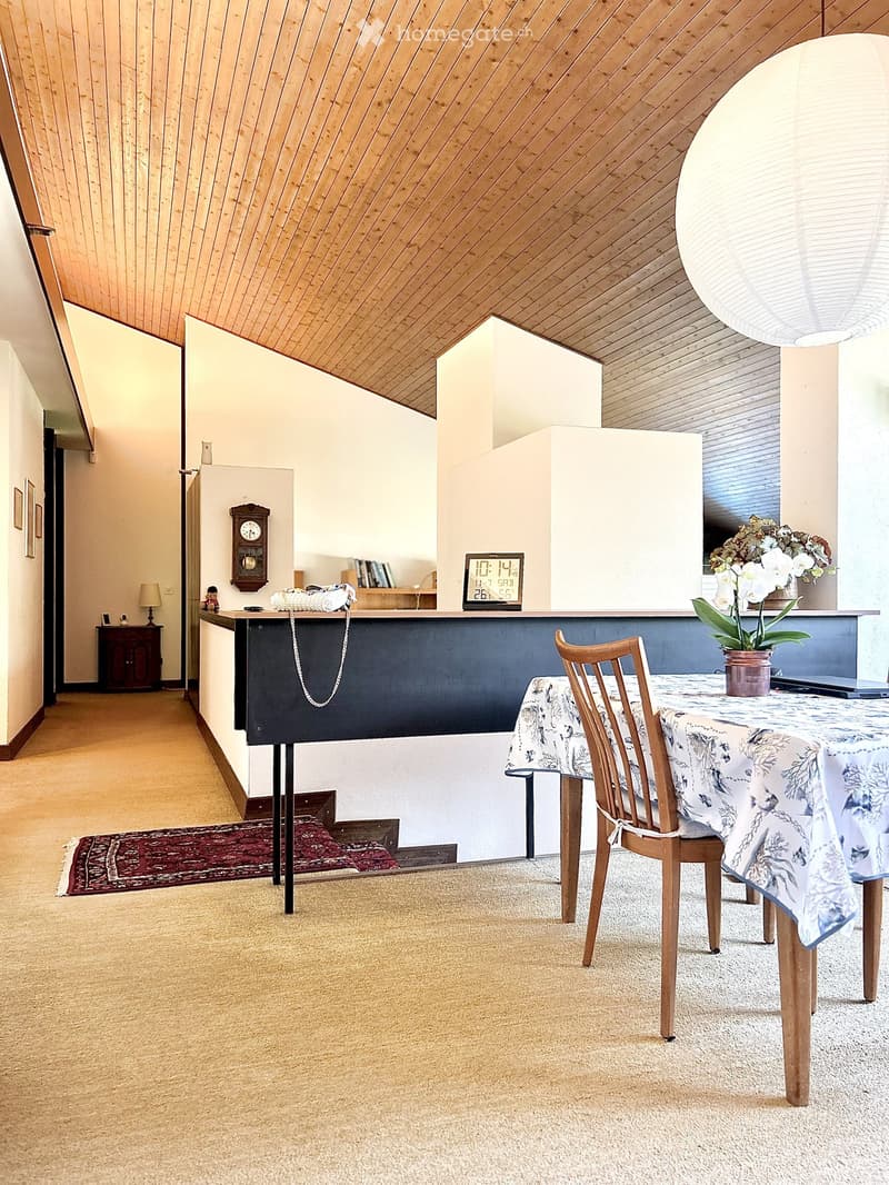 A cozy, well-lit room with a slanted wooden ceiling, a dining table with a floral tablecloth, a wooden chair, a clock on the wall, a laptop on the table, a flower vase, a small cabinet, and a lamp on a side table.