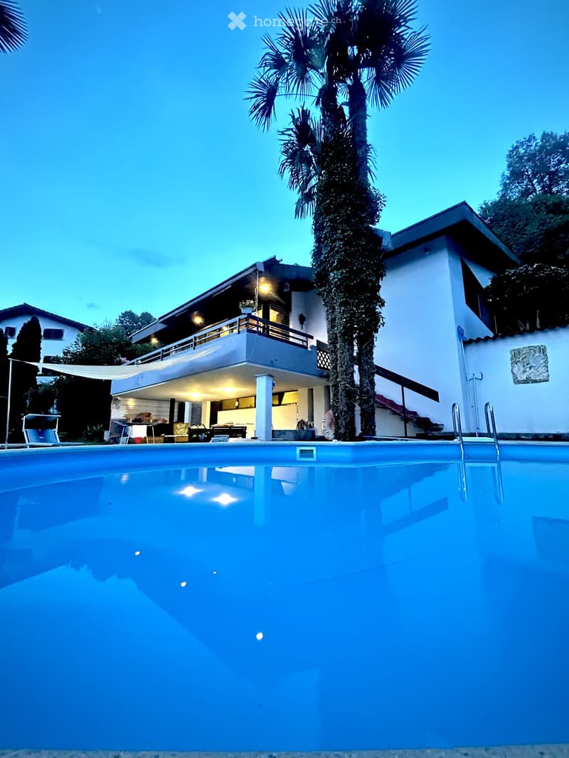 A modern two-story house with a large swimming pool in the foreground, surrounded by palm trees and lush greenery.