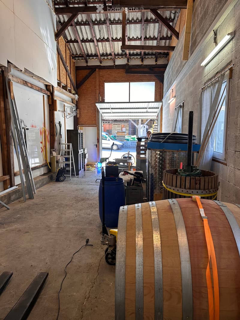 An unfurnished room with exposed wooden beams and a corrugated metal roof, featuring a large wooden barrel, a blue barrel, and various tools and equipment scattered around.
