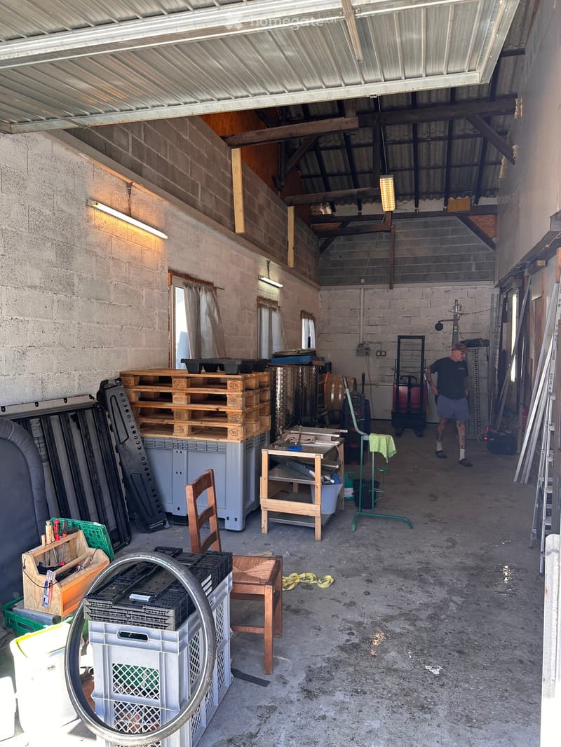 A spacious, unfinished garage with concrete floors, exposed wooden beams, and metal roofing. Contains various items including a wooden chair, a small table, stacked pallets, and a few stools.