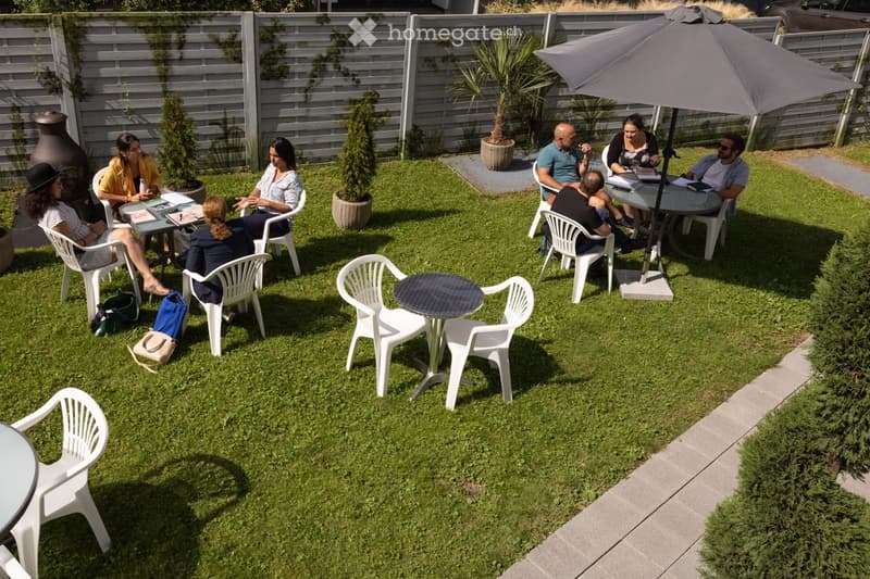 Garden area with several people seated around tables and chairs, surrounded by potted plants and a large umbrella. The setting includes a grassy lawn, a paved pathway, and a wooden fence in the background.