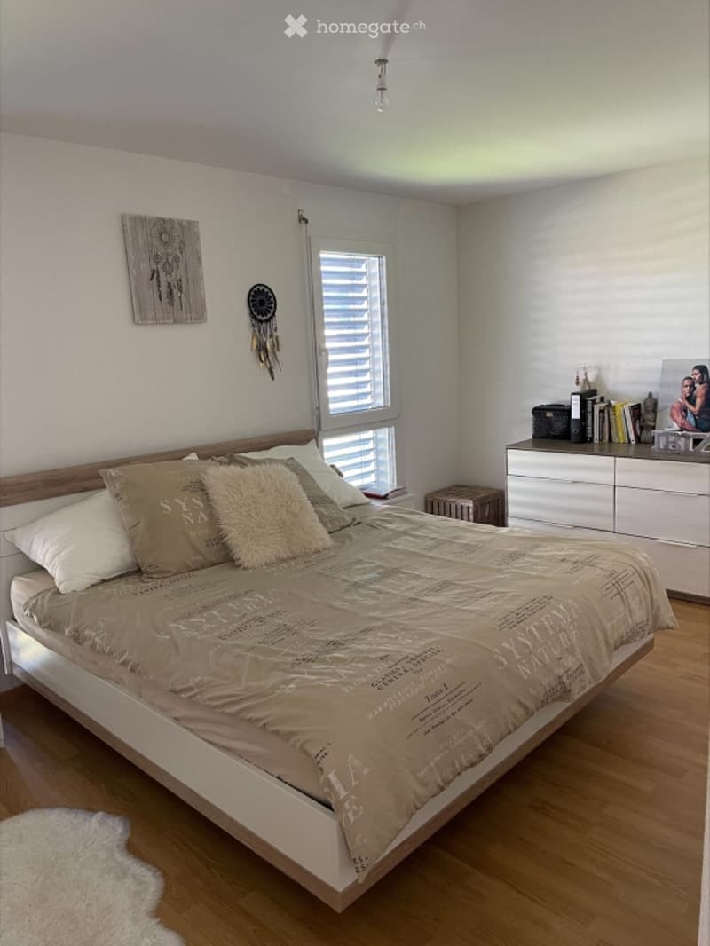 Modern bedroom with a large bed, multiple pillows, a patterned blanket, a nightstand with books, a picture frame, a dreamcatcher, and a white drawer unit.