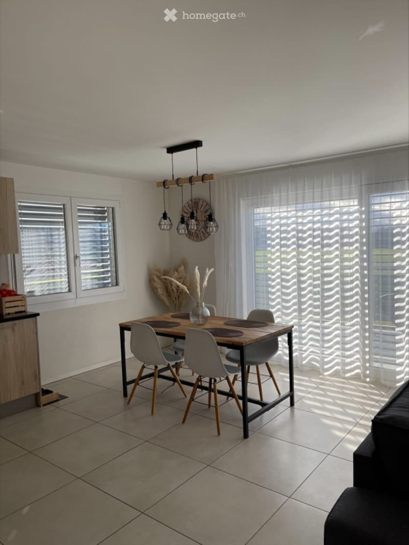 Bright dining area with a wooden dining table, four grey chairs, pendant lights, a vase with dried plants, and large windows with white curtains.