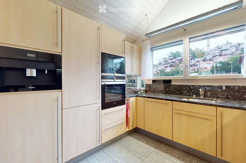 Modern kitchen with light wood cabinets, stainless steel oven, granite countertop, and a window overlooking a residential area.