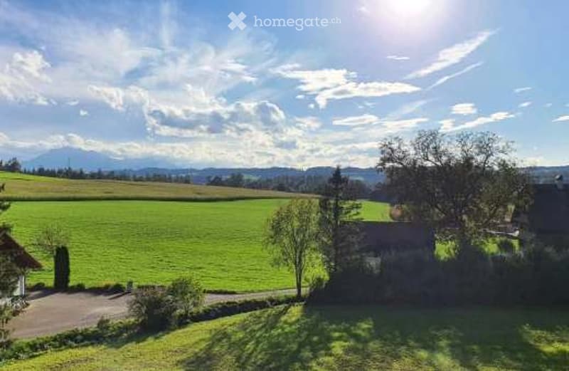 A peaceful countryside scene with green fields, scattered trees, and a small wooden structure under a partly cloudy sky.