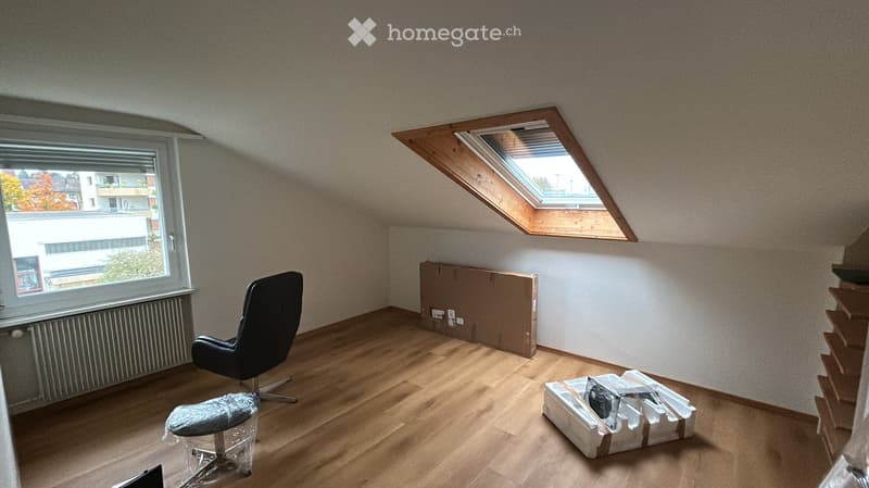 An attic room with wooden flooring, a large window, and a skylight. A black leather chair and a stool are present, along with some boxes and packaging materials.