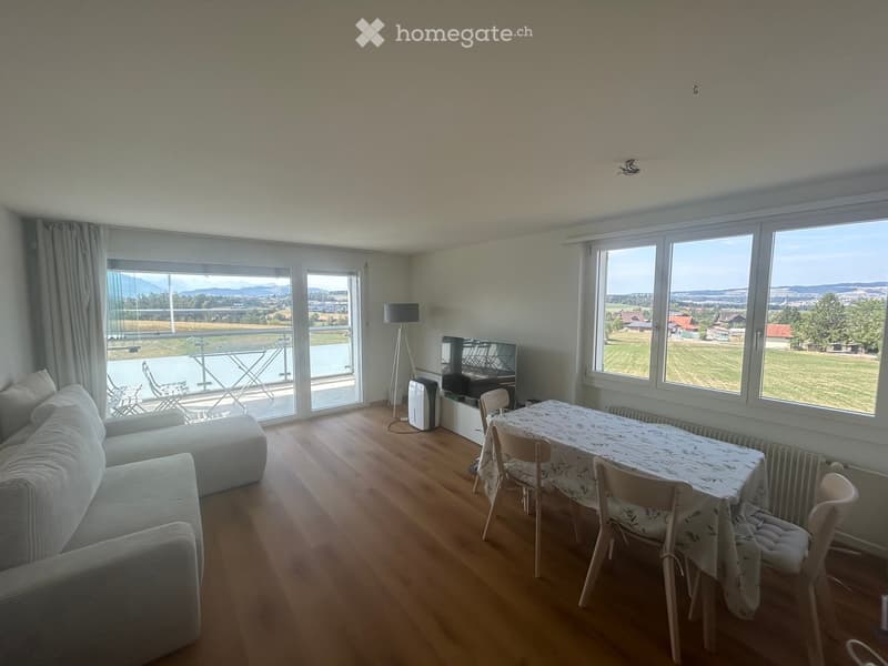 Bright living room with wooden flooring, large windows offering a scenic view, a sofa, a dining table with chairs, a television, and a floor lamp.