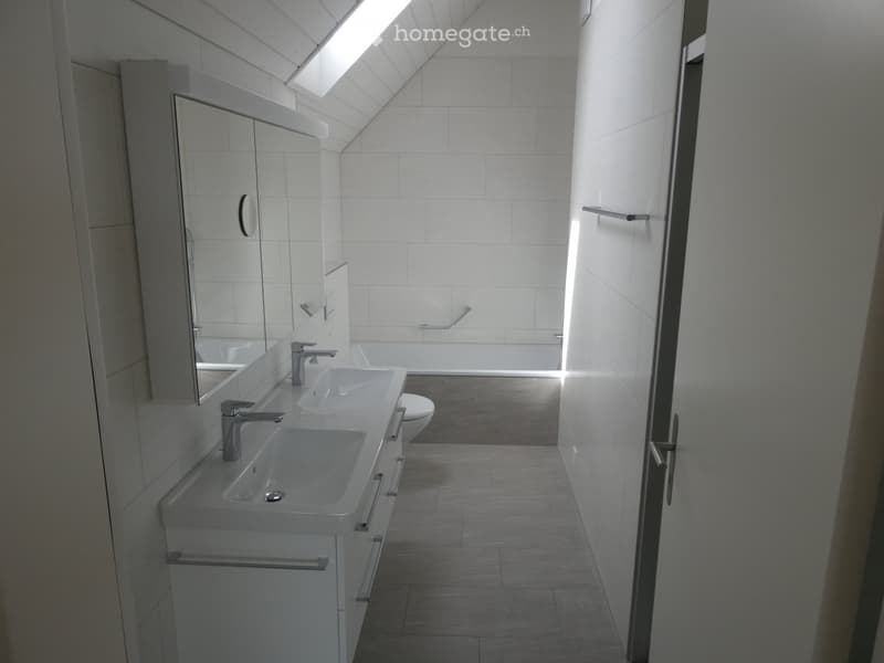 Modern bathroom with white tiles, double sink, mirror, toilet, and bathtub, featuring a skylight window for natural light.