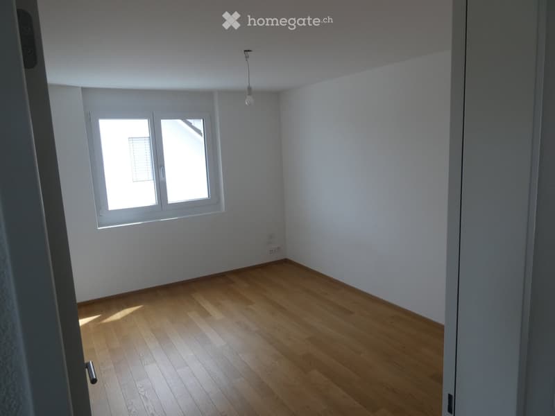 Empty room with light wooden flooring, white walls, and a window with blinds allowing natural light to enter.
