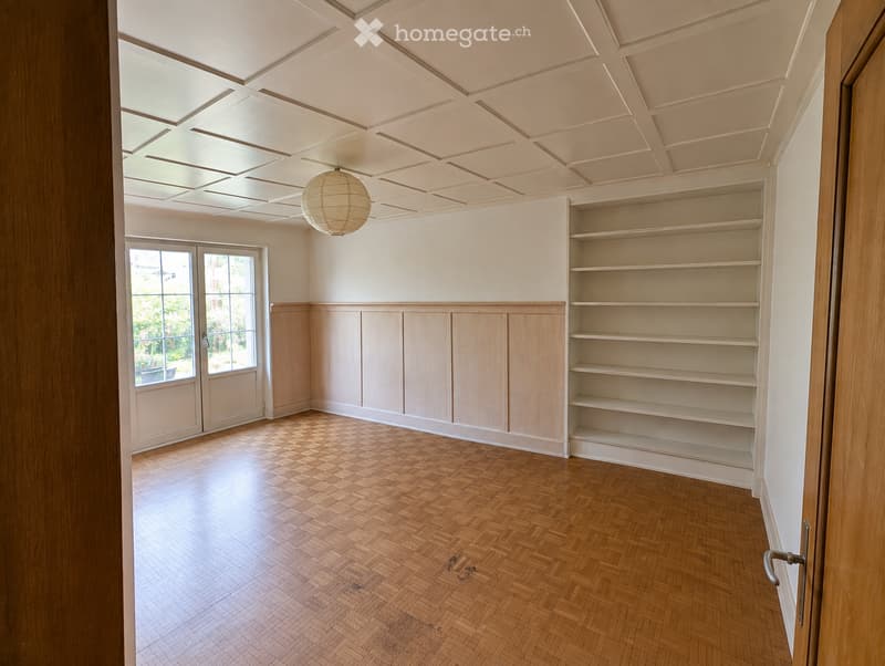 Empty room with wooden herringbone flooring, built-in wooden shelving unit, large windows, and a decorative ceiling light fixture.