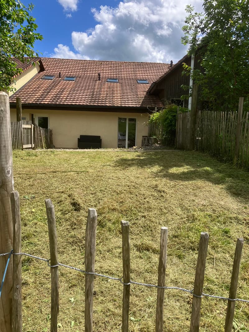 A fenced garden area with a wooden fence, grassy lawn, and a small house with a tiled roof in the background.