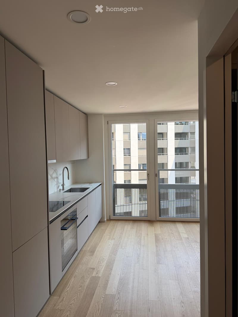 Modern kitchen with light wooden flooring, white cabinets, a sink, and an oven. Large sliding glass doors lead to a balcony with a view of neighboring buildings.
