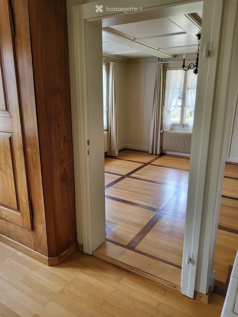 Empty room with wooden flooring, white walls, and large windows with curtains. The room is connected to a hallway with wooden doors and a chandelier visible in the adjacent space.