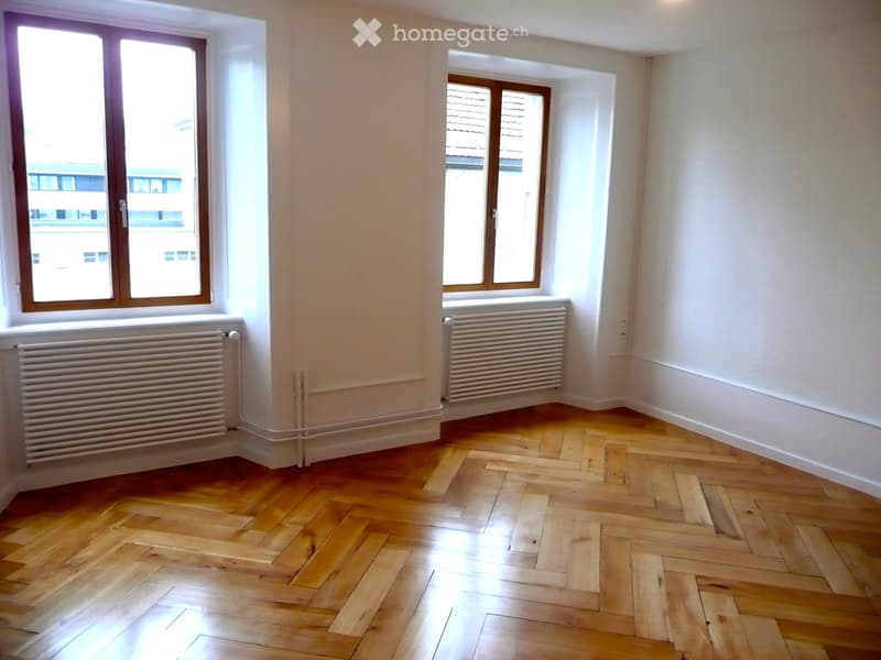 Empty room with wooden herringbone flooring, white walls, and three windows with wooden frames. Radiators are present under two of the windows.