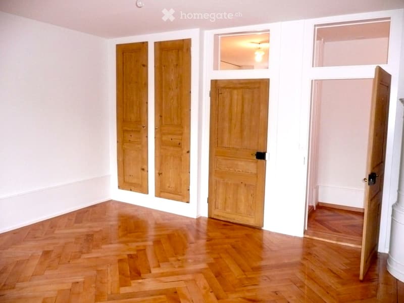Empty room with wooden herringbone flooring, white walls, and several wooden doors leading to other spaces.