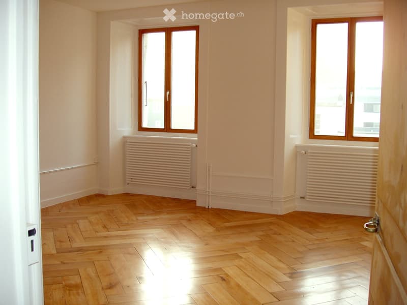 Empty room with wooden herringbone flooring, white walls, and two windows with wooden frames. Radiators are present under each window.
