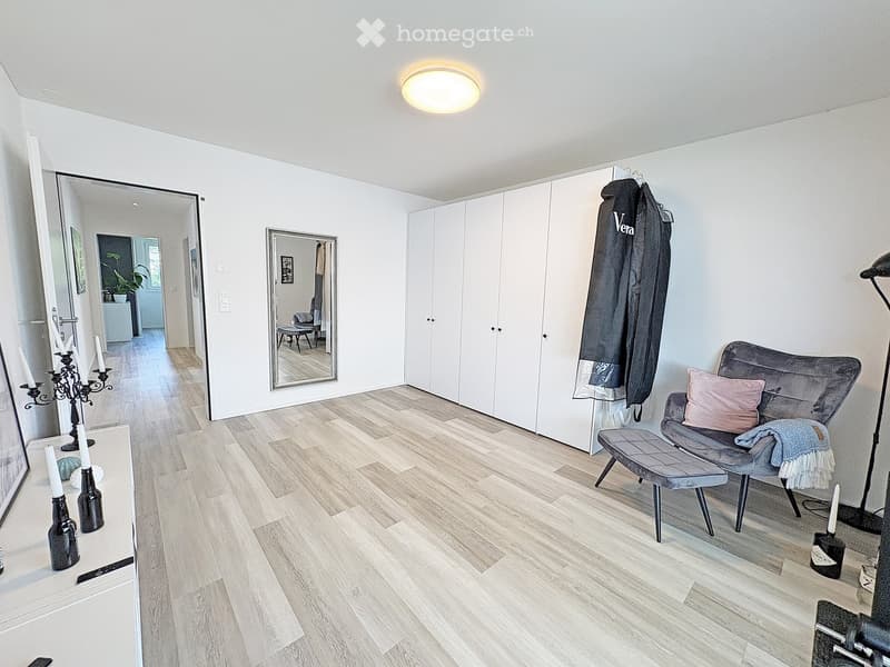Modern living room with light wooden flooring, white walls, a grey sofa with pillows, a small bench, a large mirror, and a candle holder on a side table.