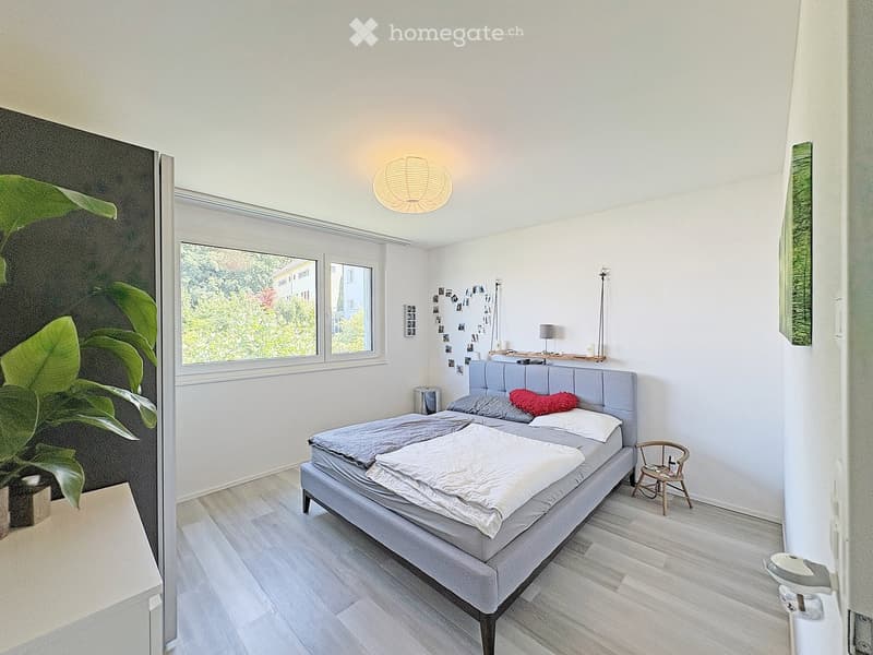 Modern bedroom with a grey upholstered bed, white bedding, a small wooden chair, a floor lamp, and a large leafy plant near the window.