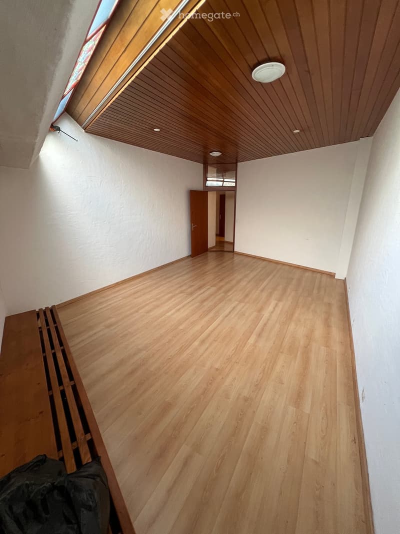 Empty room with wooden flooring, white walls, and a wooden ceiling with recessed lighting. A door leads to another room, and a window is visible at the top left corner.