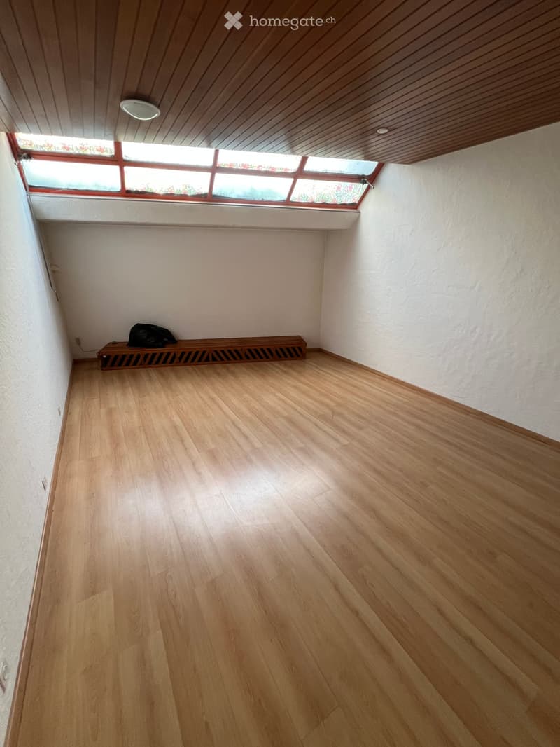 Empty room with wooden flooring, white walls, and a small bench with a bag on it. The ceiling has wooden paneling and a skylight window.