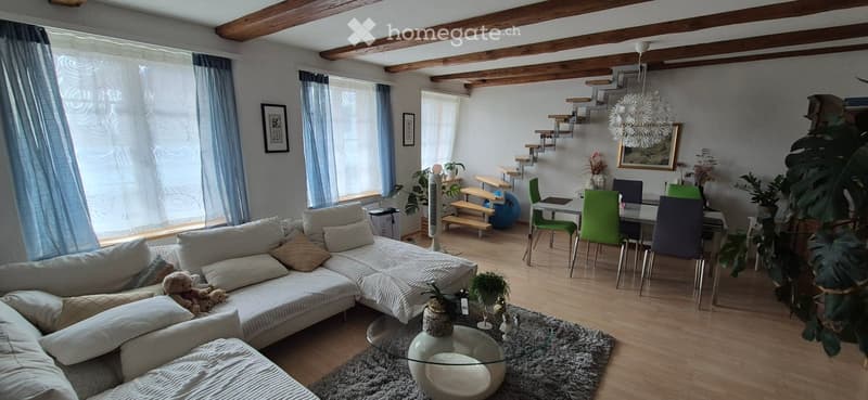 A modern living room with a large sectional sofa, glass coffee table, dining area with chairs, and decorative plants. The room features wooden beams on the ceiling and large windows with sheer curtains.