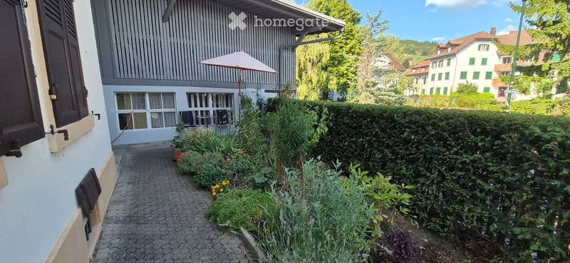A narrow garden path lined with greenery and potted plants leads to a building entrance. The building has a modern facade with large windows and a balcony area with an umbrella. The surrounding area is lush with trees and other houses visible in the backg